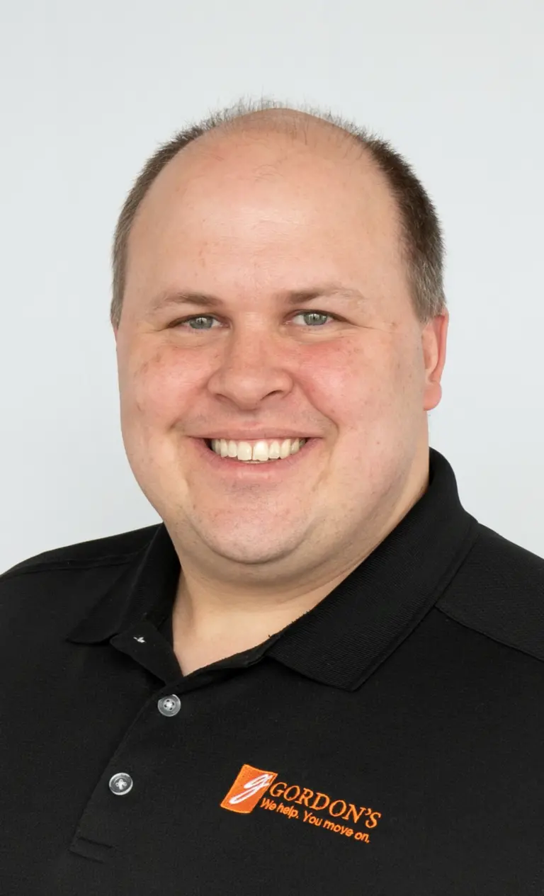 A smiling man with short hair wearing a black polo shirt with the logo "Gordon's" and the tagline "We help. You move on."