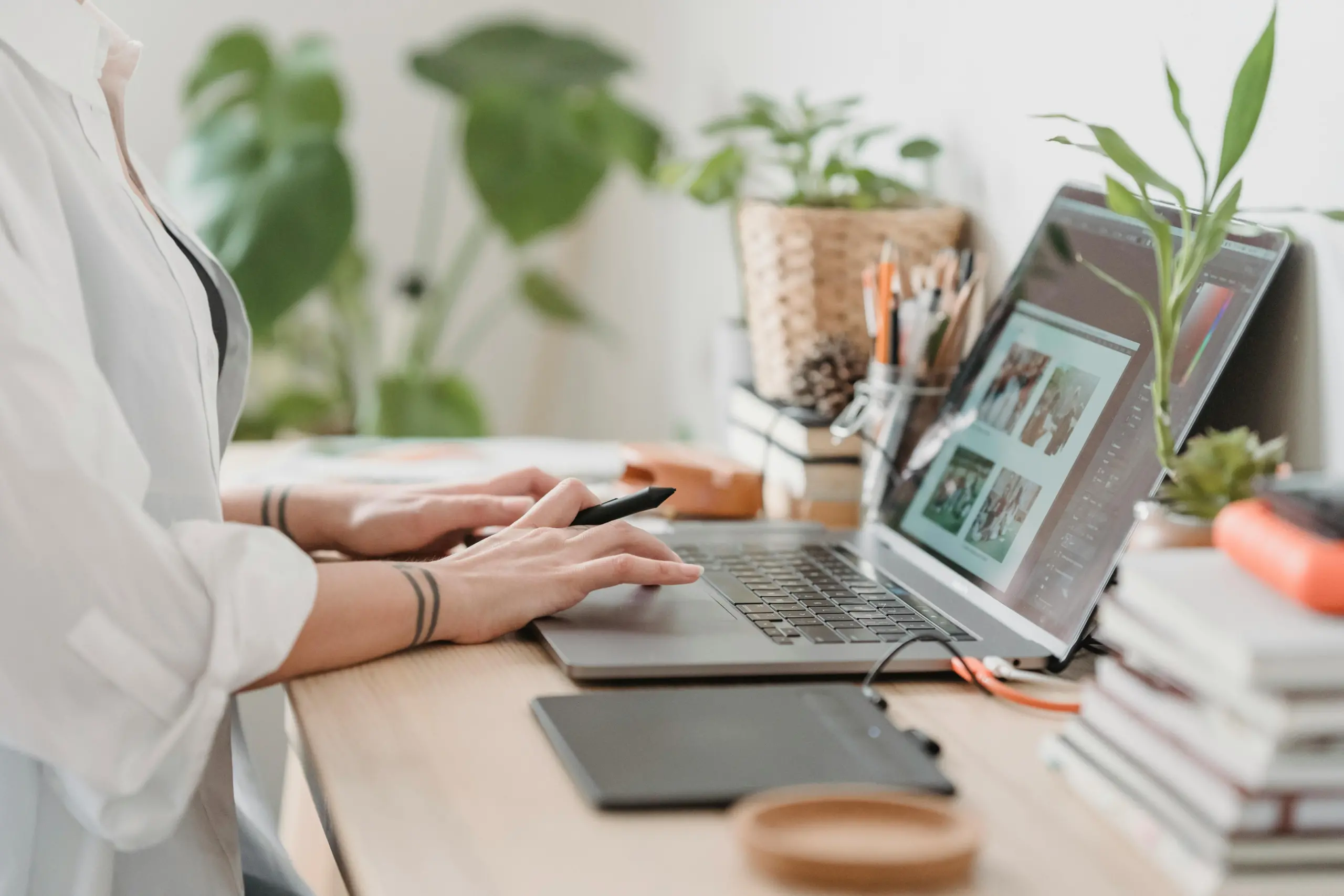 A person working on a laptop at a desk with various plants and office supplies. They are using a stylus pen and a graphics tablet, and the laptop screen displays photo editing software with images.