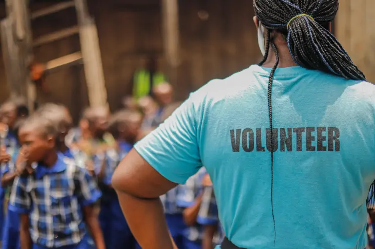 A Black woman with braided hair wearing a turquoise shirt with 'VOLUNTEER' printed on the back. She is standing in front of a group of children in blue plaid uniforms.